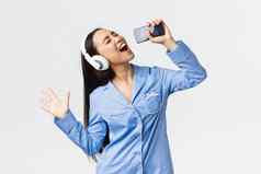 首页休闲周末生活方式概念兴奋无忧无虑的亚洲女孩睡衣玩卡拉ok应用程序智能手机唱歌首歌移动电话穿耳机白色背景