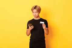 年轻的英俊的亚洲的家伙黑色的t恤购物在线持有信贷卡智能手机站黄色的背景