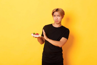 肖像恶心心烦意乱年轻的亚洲的家伙拒绝吃蛋糕不高兴显示拒绝手势黄色的背景