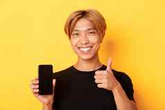 满意英俊年轻的亚洲的家伙公平头发显示竖起大拇指批准智能手机屏幕站黄色的背景