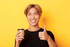 特写镜头满意亚洲英俊的的家伙推荐咖啡馆持有杯咖啡显示竖起大拇指批准微笑很高兴黄色的背景