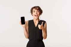 惊讶年轻的女人显示竖起大拇指移动电话屏幕推荐商店应用程序站白色背景