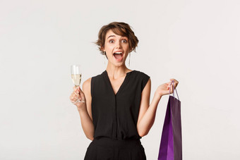 <strong>兴奋</strong>时尚的女人想知道持有购物袋玻璃香槟站白色背景庆祝活动<strong>聚会</strong>，派对