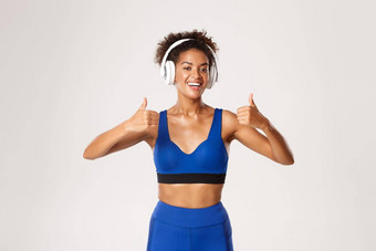 健康的运动非裔美国人女人蓝色的运动服装显示竖起大拇指听音乐耳机练习锻炼白色背景