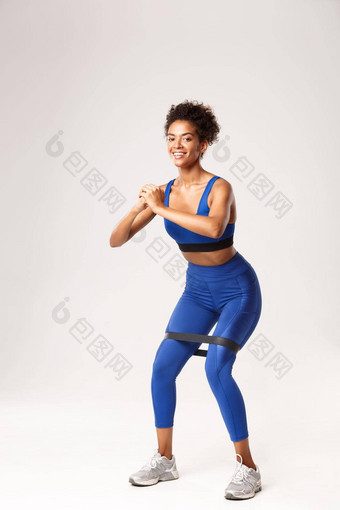 完整的长度微笑适合<strong>女人</strong>享受健身穿蓝色的体育运动装<strong>下蹲</strong>电阻乐队站白色背景
