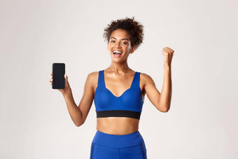 <strong>体育运动</strong>技术概念快乐的非裔美国人健身女人显示锻炼应用程序智能<strong>手机</strong>屏幕使拳头泵并实现每天目标白色背景