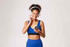 美丽的健身女人蓝色的运动服装锻炼音乐无线耳机智能手机应用程序站白色背景