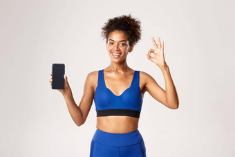 体育运动技术概念满意非裔美国人女运动员蓝色的健身服装显示智能手机屏幕标志白色背景