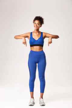 完整的长度快乐非裔美国人健身教练蓝色的运动服装指出手指微笑推荐锻炼齿轮显示标志体育促销白色背景
