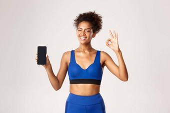 <strong>体育运动</strong>技术概念有吸引力的健身女人蓝色的锻炼服装显示智能<strong>手机</strong>屏幕标志眨眼微笑推荐应用程序