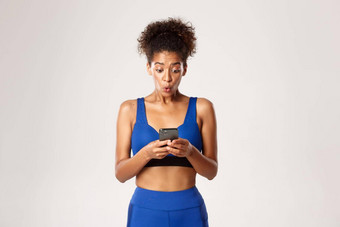 体育运动技术概念惊讶非裔美国人适合女孩锻炼服装兴奋智能手机屏幕站白色背景