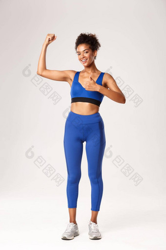 完整的长度强大的有吸引力的非洲美国女运动员蓝色的运动服装显示竖起大拇指flex肱二头肌推荐健身房锻炼好培训会话白色背景