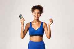 快乐的非裔美国人女运动员体育运动装欢乐微笑持有智能手机信贷卡站白色背景