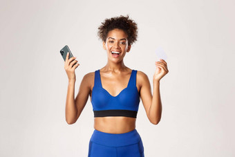 快乐的非裔美国人女运动员<strong>体育运动</strong>装欢乐微笑持有智能<strong>手机</strong>信贷卡站白色背景