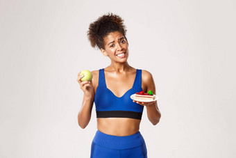 健康的生活方式体育运动概念悲观的非裔美国人女运动员饮食持有蛋糕绿色苹果<strong>抵制</strong>诱惑白色背景