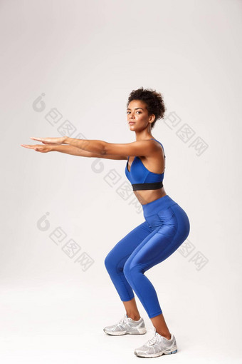 完整的长度美丽的健康的非裔美国人女人蓝色的体育运动服装伸展运动手下蹲练习相机worout白色背景