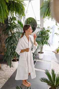 完整的长度拍摄快乐的年轻的女人白色浴袍微笑应用唇乳香嘴唇站浴室装饰植物