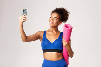 年轻的美丽的非裔美国人健身女人<strong>体育运动</strong>装采取自拍瑜伽席采取照片智能<strong>手机</strong>站白色背景