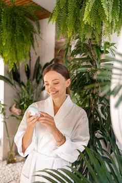快乐年轻的女人浴袍持有硅胶刷清洗设备美例程浴室装饰植物