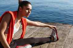 户外拍摄有吸引力的健身女人准备活动慢跑坐着码头伸展运动腿