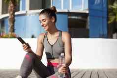户外拍摄快乐健身女人移动电话应用程序坐着木地板上喝水