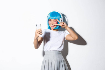 肖像可爱的亚洲女孩穿着动漫字符摆姿势自拍耳机蓝色的<strong>短假发</strong>采取图片智能手机站白色背景