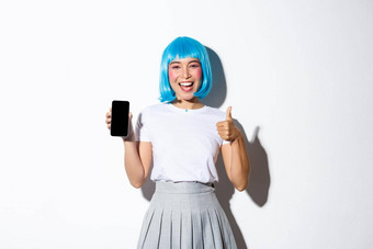 肖像快乐微笑亚洲女孩蓝色的短假发显示翘拇指批准智能手机应用程序显示移动电话屏幕站白色背景