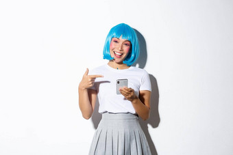 肖像美丽的亚洲女孩穿着动漫字符蓝色的短假发指出手指智能手机微笑站白色背景