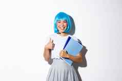 图像可爱的微笑亚洲女孩穿蓝色的动漫假发持有学校笔记本电脑穿着女学生万圣节聚会，派对显示竖起大拇指批准满意