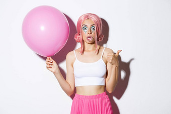 图像兴奋有吸引力的女孩吹泡沫口香糖显示竖起大拇指批准站可爱的粉红色的气球穿聚会，派对装假发