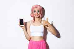 图像有吸引力的年轻的聚会，派对女孩粉红色的假发明亮的化妆显示竖起大拇指移动电话屏幕推荐应用程序站白色背景