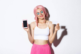 肖像可爱的很高兴女孩快乐关闭眼睛微笑显示信贷卡智能手机屏幕站粉红色的动漫假发明亮的化妆