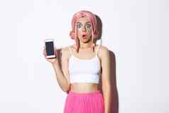 图像惊讶女孩敬畏显示智能手机屏幕穿着粉红色的假发聚会，派对装站白色背景