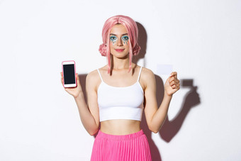 图像时尚的聚会，派对女孩粉红色的假发显示信贷卡移动电话屏幕庆祝假期魅力装站白色背景