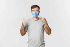 概念流感大流行科维德社会距离图像快乐的家伙灰色的t恤指出医疗面具显示竖起大拇指站白色背景