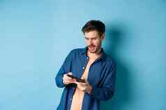 年轻的的家伙玩视频游戏移动电话倾斜身体持有智能手机水平站蓝色的背景