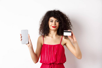在线购物时尚女人卷曲的头发穿红色的衣服显示智能手机屏幕塑料信贷卡微笑相机白色<strong>背景</strong>