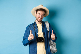 英俊的高加索人旅游稻草他持有背包照片相机显示拇指推荐旅行机构酒店度假胜地蓝色的背景