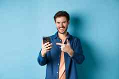 英俊的微笑男人。指出手指智能手机很高兴相机推荐应用程序站蓝色的背景