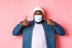 冠状病毒生活方式全球流感大流行概念年轻的非裔美国人男人。指出脸面具显示竖起大拇指保护科维德粉红色的背景