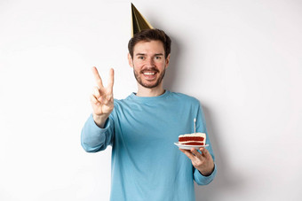 庆祝活动假期概念快乐年轻的男人。庆祝生日采取图片和平标志穿聚会，派对他持有bday蛋糕白色背景