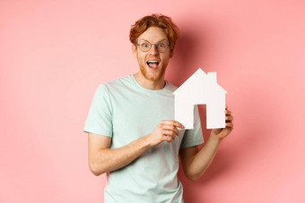 真正的房地产快乐的红色头发的人男人。购买财产显示纸房子断路微笑惊讶站粉红色的背景