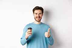 快乐的年轻的男人。喝好咖啡持有纸杯显示拇指推荐咖啡馆商店站白色背景