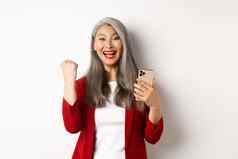 亚洲女人赢得在线持有智能手机使拳头泵手势庆祝赢得并微笑站白色背景