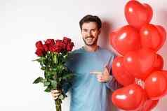 情人节一天浪漫快乐年轻的男人。花心气球日期情人微笑的家伙指出花束红色的玫瑰站白色背景