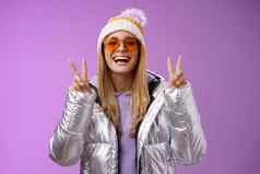 友好的幸运的乐观美丽的金发碧眼的女人太阳镜银时尚的夹克冬天他显示和平胜利手势享受雪假期酒店享受滑雪紫色的背景