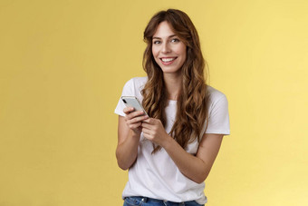 热情的好看的城市女孩穿白色t恤站休闲微笑很高兴相机发短信持有智能手机滚动社会媒体饲料站黄色的背景浏览网络