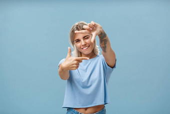 有创意的快乐肖像迷人的热情的女设计师金发碧眼的头发纹身手臂眨眼微笑快乐使框架手势测量空间蓝色的背景