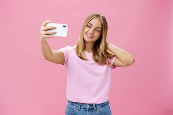 女美博主采取自拍帖子在线肖像迷人的晒黑年轻的女人t恤触碰头发轻轻拉手智能手机脸采取照片粉红色的墙
