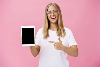 女自由<strong>程序</strong>员自豪地显示应用<strong>程序</strong>数字平板电脑持有<strong>小</strong>工具指出设备屏幕微笑广泛的很高兴表达式穿眼镜摆姿势粉红色的墙